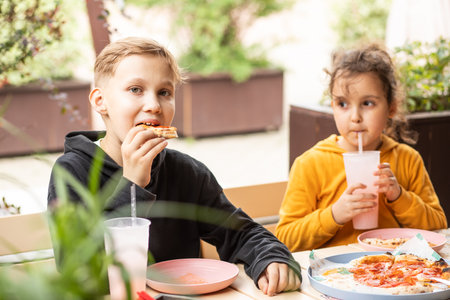 Cute girl and teenager boy brother and sister eat pizza lunch and drinking lemonade in outdoor cafe and having fun. Happy family.の写真素材