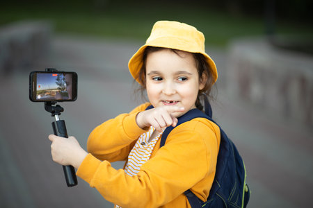 Cute little girl in yellow hat taking selfie with mobile phone outdoor in a park. Kid blogger recording video vlog.の写真素材