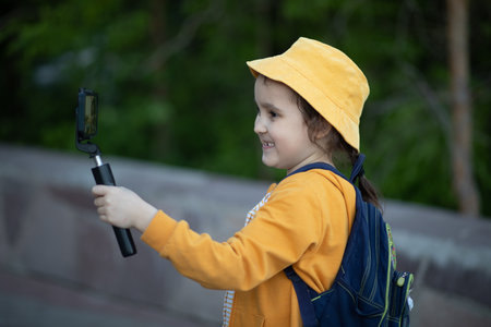 Cute little girl in yellow hat taking selfie with mobile phone outdoor in a park. Kid blogger recording video vlog.の写真素材