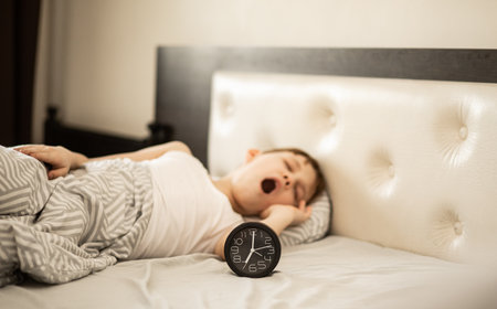 Kid boy teenager waking up morning and yawning. Alarm clock nearbyの写真素材