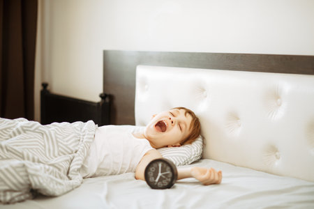Kid boy teenager waking up morning and yawning. Alarm clock nearbyの写真素材