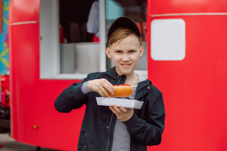 Cute teen boy kid eating hot dog nearby food track on a street. Food delivery or take away food conceptの写真素材