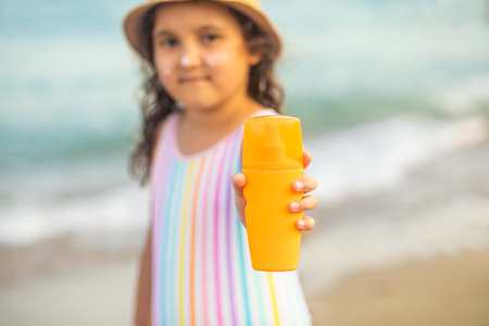 Cute little girl in a hat, striped swimsuit holding bottle of sunscreen in a hand at the beach in summer nearby sea. Holiday vacation with childrenの写真素材