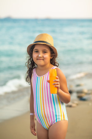 Cute little girl in a hat, striped swimsuit holding bottle of sunscreen in a hand at the beach in summer. Holiday vacation with childrenの写真素材
