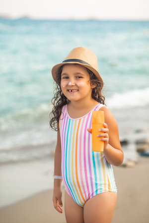 Cute little girl in a hat, striped swimsuit holding bottle of sunscreen in a hand at the beach in summer. Holiday vacation with childrenの写真素材