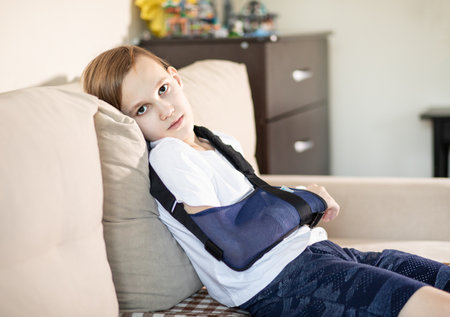 Sad kid teenager boy with a cast and bandage sitting on a sofa at home. Broken thumb finger.の写真素材