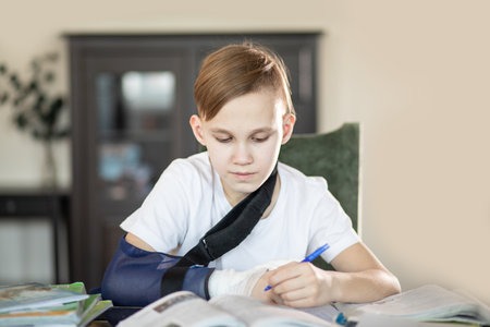 Kid boy pupil at home making homework with broken finger. Teenager hand with a cast. Hand wrapped in bandage.の写真素材
