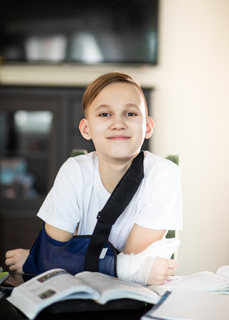 Kid boy pupil at home making homework with broken finger. Teenager hand with a cast. Hand wrapped in bandage.の写真素材