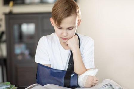 Kid boy pupil at home making homework with broken finger. Teenager hand with a cast. Hand wrapped in bandage. Concept of child insurance and healthcare.の写真素材