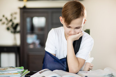 Kid boy pupil at home making homework with broken finger. Teenager hand with a cast. Hand wrapped in bandage.の写真素材