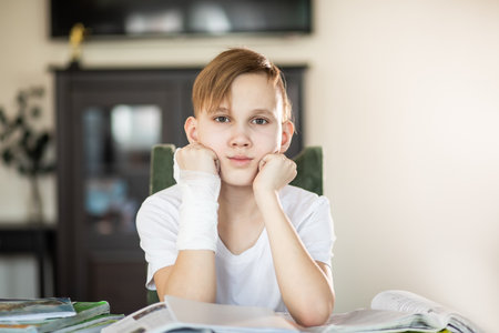 Kid boy pupil at home making homework with broken finger. Teenager hand with a cast. Hand wrapped in bandage. Concept of child insurance and healthcare.の写真素材