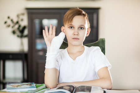 Kid boy pupil at home making homework with broken finger. Teenager hand with a cast. Hand wrapped in bandage. Concept of child insurance and healthcare.の写真素材