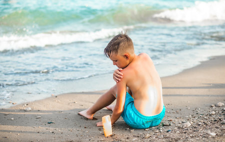 Cute little kid boy applying sunscreen at the beach in summer sitting on the seashore nearby sea. Family holiday vacation with childrenの写真素材