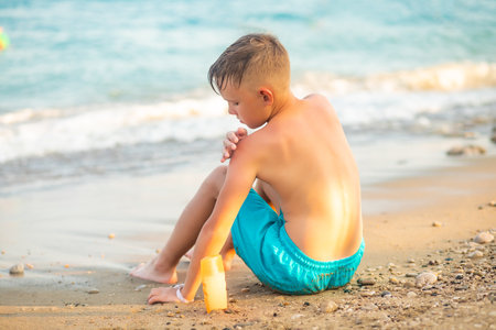 Cute little kid boy applying sunscreen at the beach in summer sitting on the seashore nearby sea. Family holiday vacation with childrenの写真素材