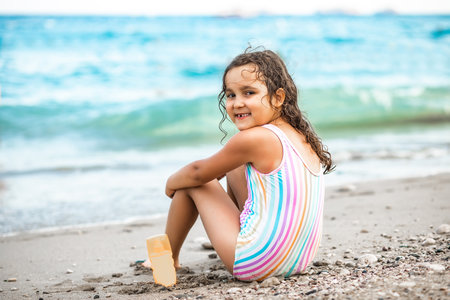 Cute little girl in striped swimwear sitting on the seashore nearby sea at the beach in summer. Playful active kid. Holiday vacation with childrenの写真素材