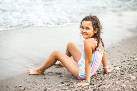Cute little girl in striped swimwear sitting on the seashore nearby sea at the beach in summer. Playful active kid. Holiday vacation with childrenの写真素材