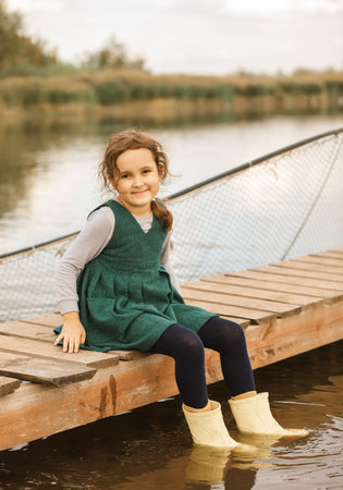 Cute little girl sits on a wooden pier by the lake, river or pond. Autumn season.の写真素材