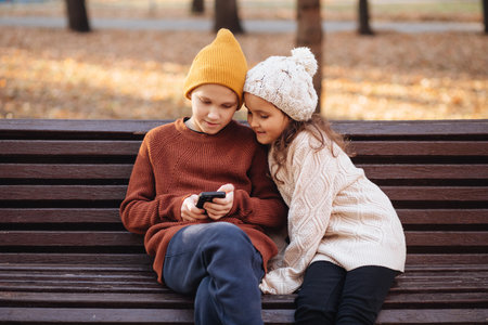 Kids girl and boy play online games or read social networks on mobile smart phones. Children sitting on bench outdoor in park.の写真素材