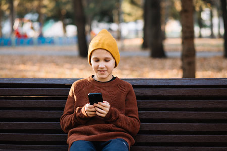 Kid boy teenager play online games or read social networks on mobile smart phones. Child sitting on bench outdoor in park.の写真素材