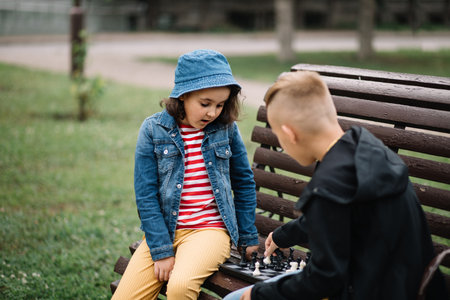 Kids girl and teenager boy playing chess outdoor in a summer park sitting on benchの写真素材