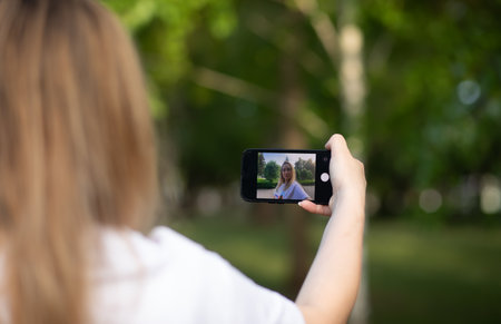 Pretty woman taking selfie with mobile phone looking at camera in a park. Blogger recording video...