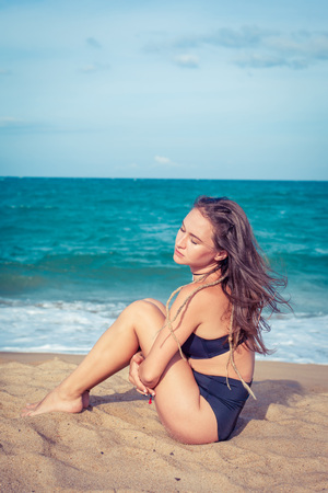 Sexy woman with tan skin in a black swimsuit sitting on the sand of the beach. Female hug her knees near sea.の写真素材
