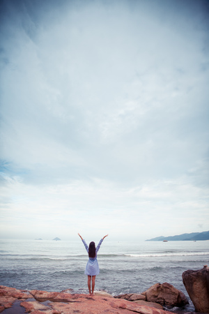 Asian woman in dress back stand and rise hands on a stone by the seaの写真素材