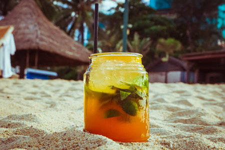 Close up of glass with refreshing orange juice tcocktail with lime and grass on sand of tropical beachの写真素材