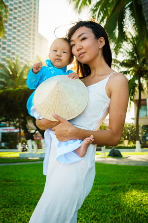 A mother holds her little daughter in traditional vietnam clothers aodai in her arms on the beach near palmsの写真素材