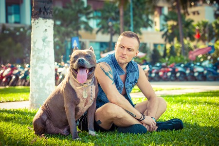 Happy friends man and dog american pit bull terrier sitting on grass in parkの写真素材