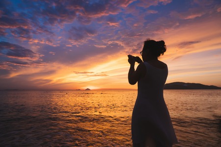 Woman in dress make photo of beautiful colorful sunrise over the ocean on smartphone.の写真素材