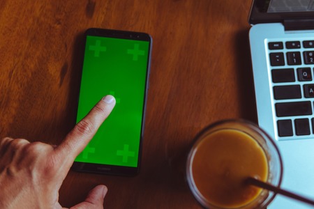 Finger of man tap on smartphone with green screen near. Iced coffee and laptop on a wooden table