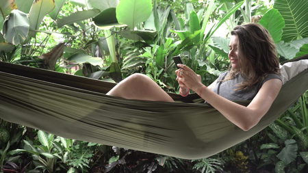 Woman using smartphone while sitting on hammock, exotic backgroundの写真素材
