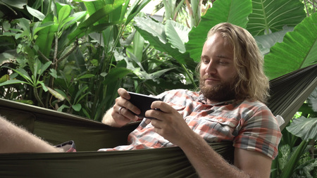 Young man playing game on mobile phone while sitting on hammockの写真素材