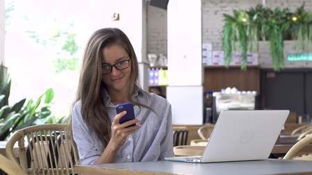 Woman in eyeglasses using her smartphone, laptop on the tableの写真素材