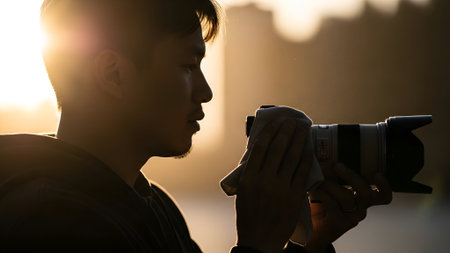 A focused photographer, silhouetted against the warm golden hour sun, holds a professional camera with a telephoto lens outdoors, ready to capture the perfect shot.の素材
