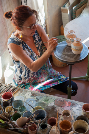 The craftsman paints two small ceramic pots in his shop. Various brushes and colours on his bench.の写真素材