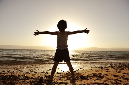 Silhouette of child on the beach, holding his hands up, towards the sunの写真素材