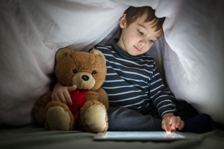 Cute little kid with his friend teddy bear using tablet computer under blanket at night in a dark roomの写真素材