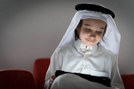 Cute Arabian kid sitting on sofa at home with tabletの写真素材