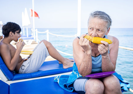Senior woman sitting on beach pier and eating cornの写真素材