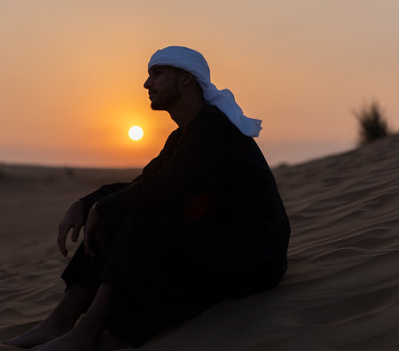 Arab man sitting on sand in the desert during golden hour, wearing traditional arab clothing. High quality photoの写真素材