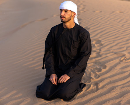 Arab man sitting on sand in the desert during golden hour, wearing traditional arab clothing. High quality photoの写真素材
