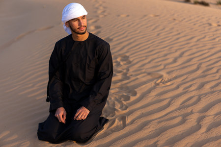 Arab man sitting on sand in the desert during golden hour, wearing traditional arab clothing. High quality photoの写真素材