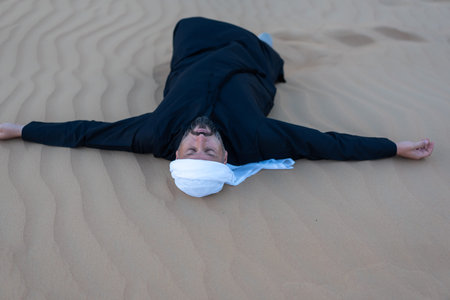 Relaxing male tourist wearing traditional arab clothing, lying on desert sand dunes. High quality...