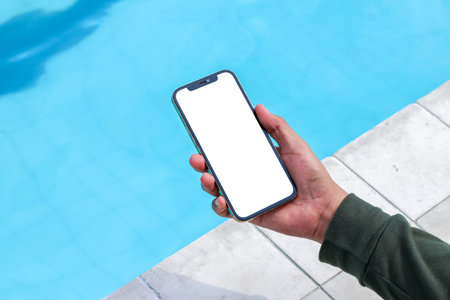 Image of a man browsing on to a white blank smartphone near a pool for mockupの写真素材
