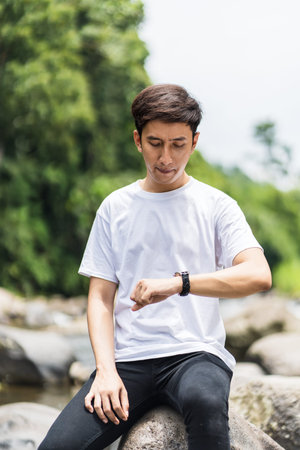 A sitting young asian man with blank white shirt checking on his watch with a river as the...