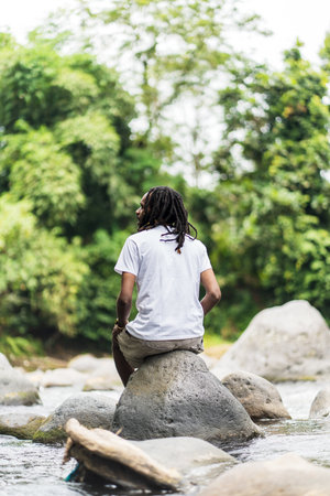 Back side of sitting young african man with blank white shirt with a river as the background in the natureの写真素材