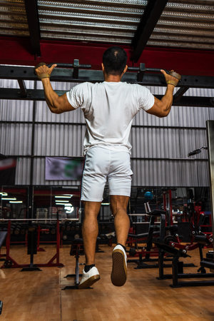Back side of a gym athlete wearing a white blank shirt doing a pull up at a gym, mockup image