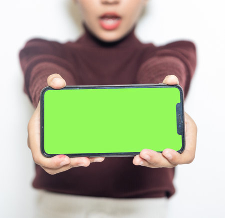 A woman holding a smartphone with green colored screen while surprised on the white backgroundの写真素材